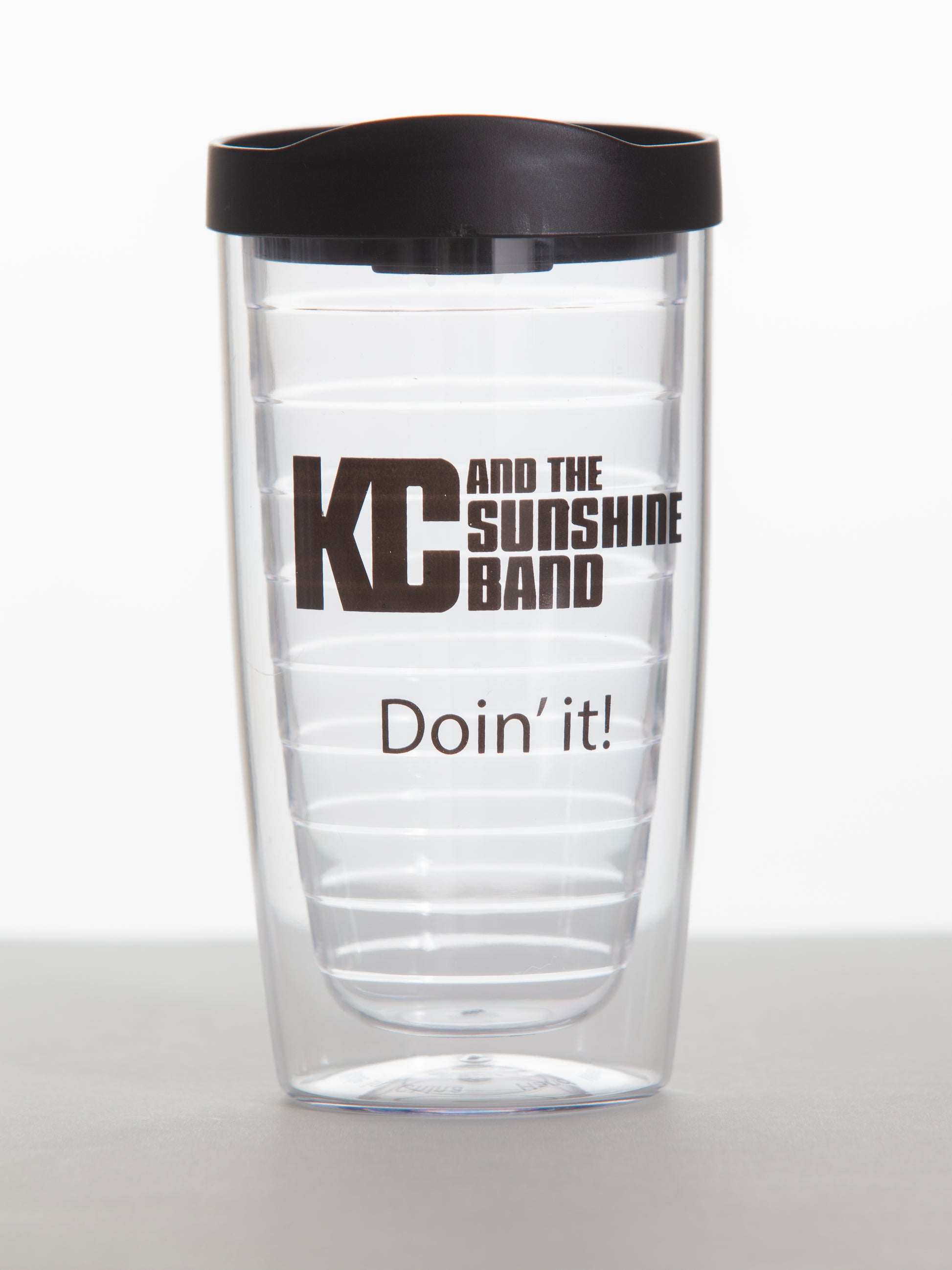 A clear plastic tumbler with a black lid, featuring the text 'KC and the Sunshine Band' and 'Doin' it!' printed in black.